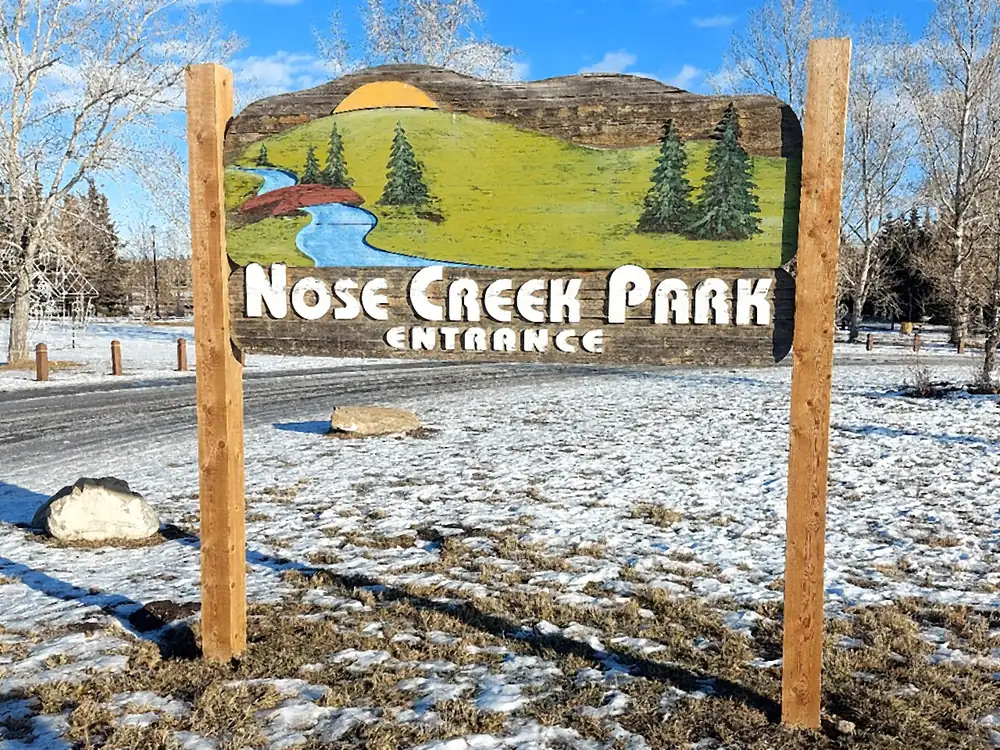 The painted wooden Nose Creek Park entrance sign in Airdrie, standing in a light dusting of snow