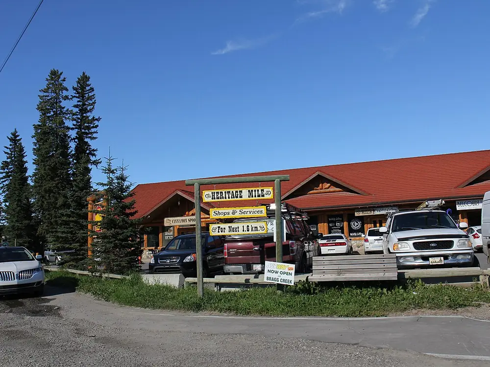 A log-style row of shops in Bragg Creek with a red roof, tall spruce trees and a wooden sign